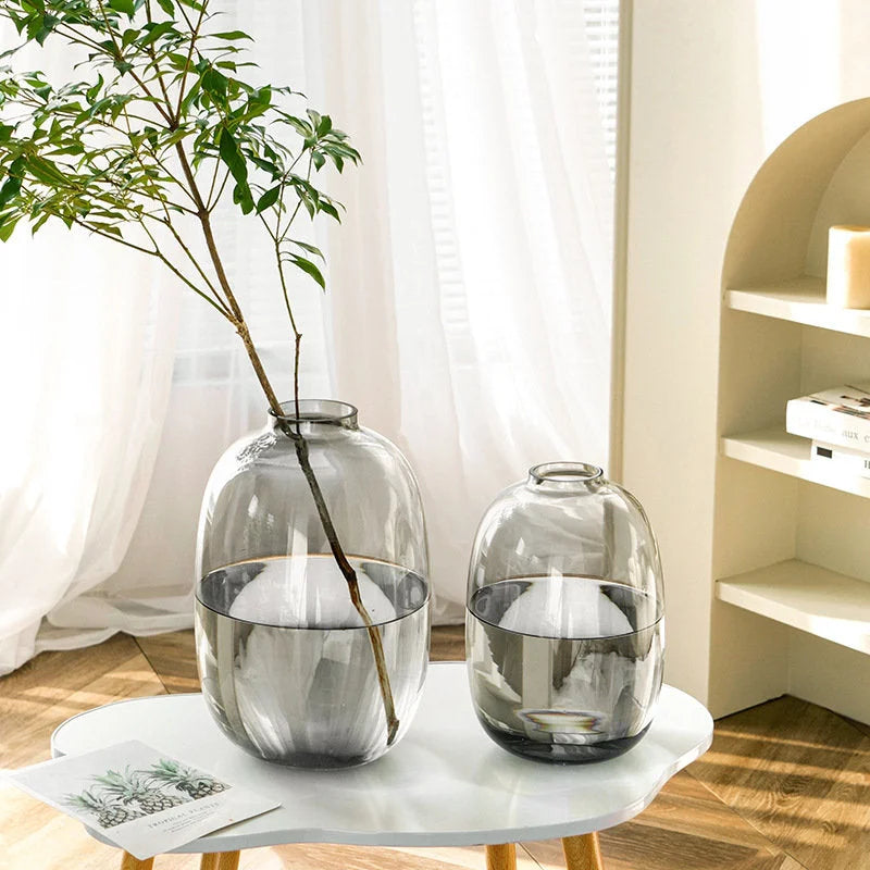 Lifestyle shot of two smoky grey glass vases on a modern white coffee table in a bright living room. - RoomDen