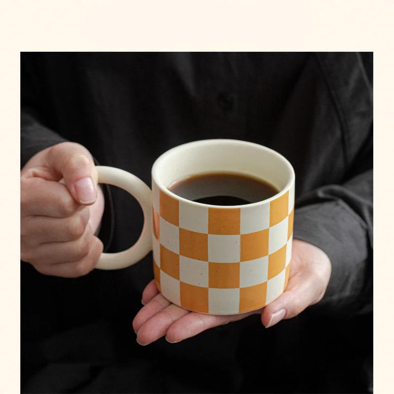 A person holding the Orange Checkered Ceramic Mug with a Puffy Handle, filled with coffee. - RoomDen