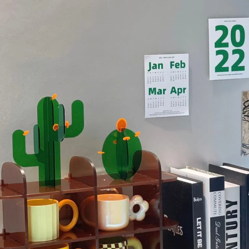 Green acrylic cactus decor styled on a brown bookshelf with mugs, books, and a 2022 calendar - RoomDen