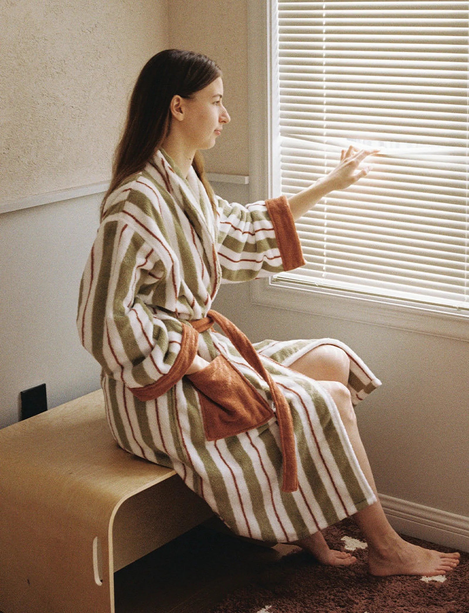 Model looking through window blinds wearing the sage green and rust striped cotton robe.