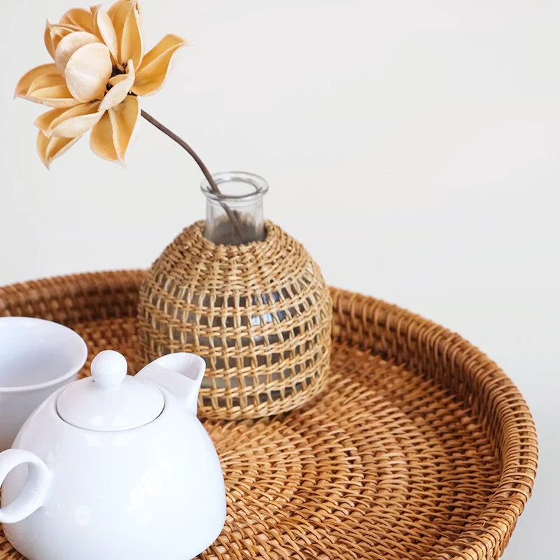 A Handwoven Rattan & Glass Bud Vase with a flower, styled on a round rattan tray with a white teapot.