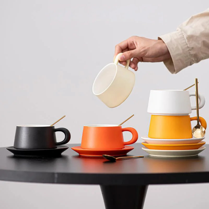 A collection of Matte Ceramic Coffee Cups and Saucers in black, orange, yellow, white, and beige on a black table. - RoomDen