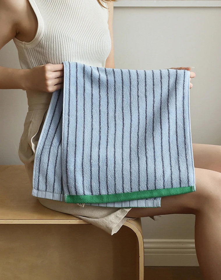 Woman holding a folded light blue pinstripe cotton face towel, showing the texture and green border detail - RoomDen