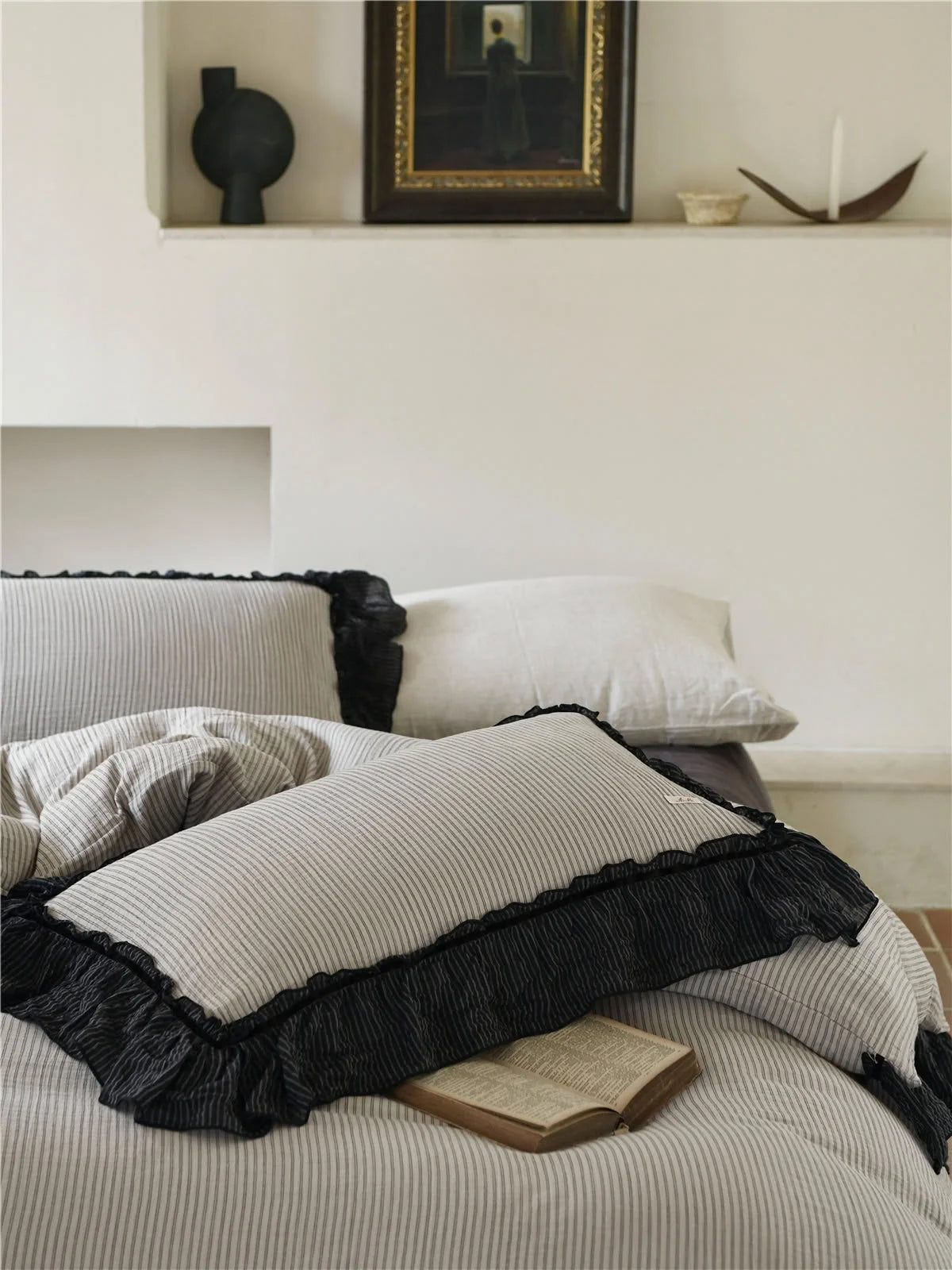 Close-up of the pinstripe pillows with black ruffle trim, with a book resting on them and art on a shelf behind. - RoomDen