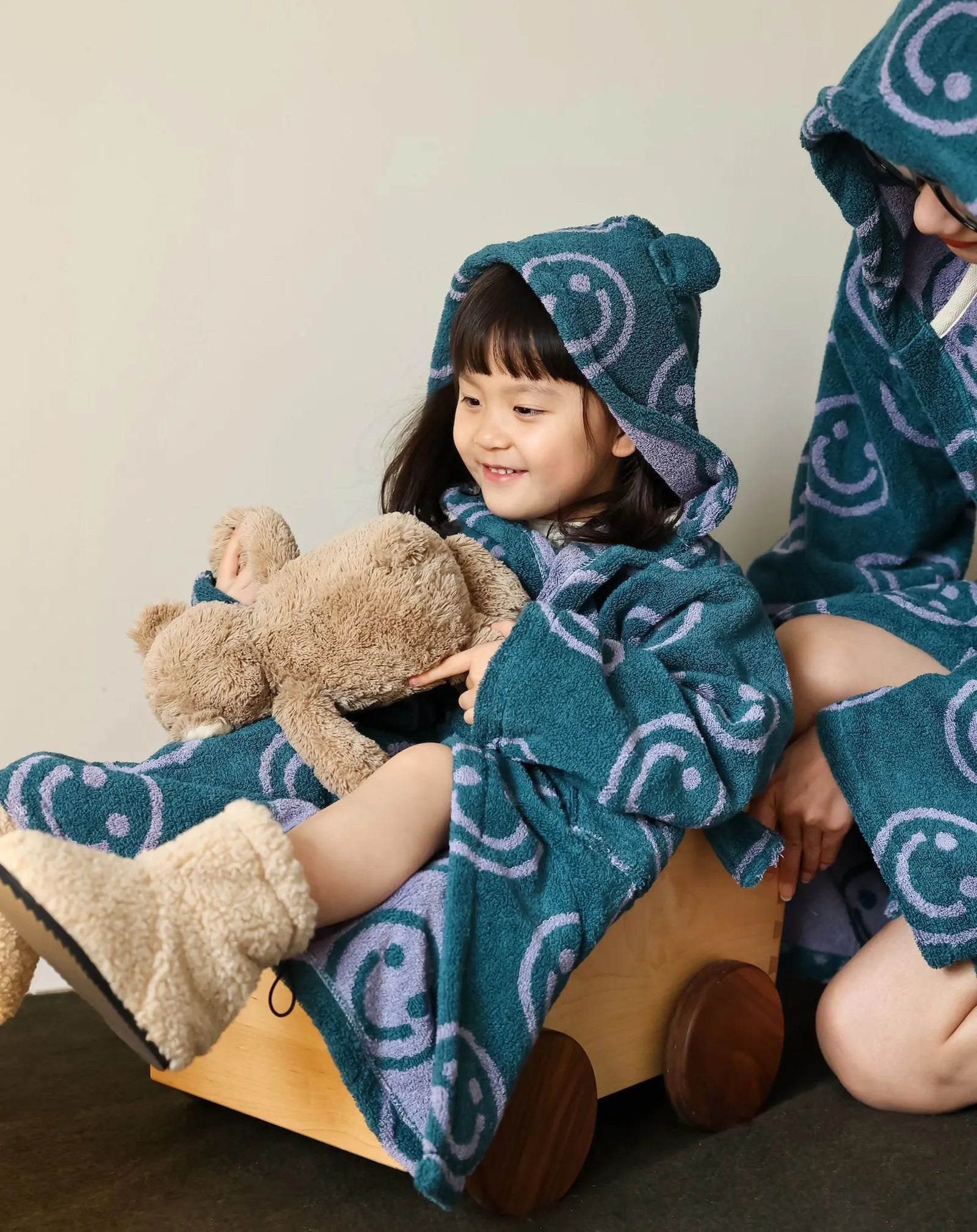 Close up of a child wearing a teal and purple smiley face terry cloth robe holding a beige teddy bear