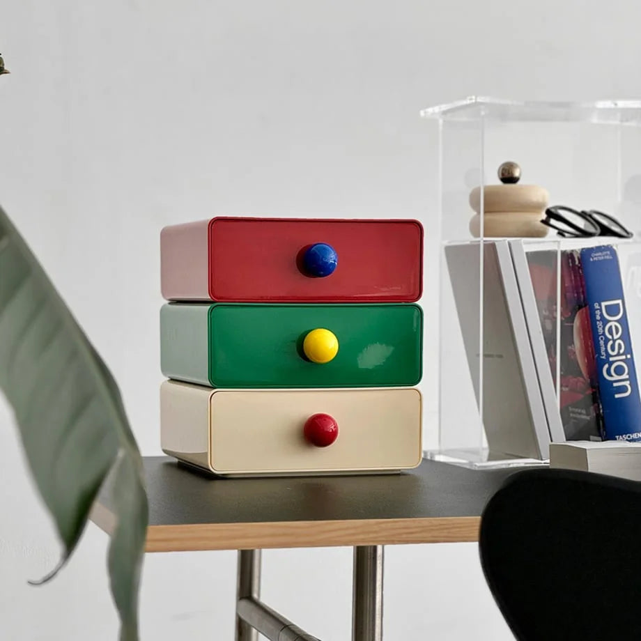 A set of three stacked desk drawers in red, green, and beige on a modern desk with a plant. - RoomDen