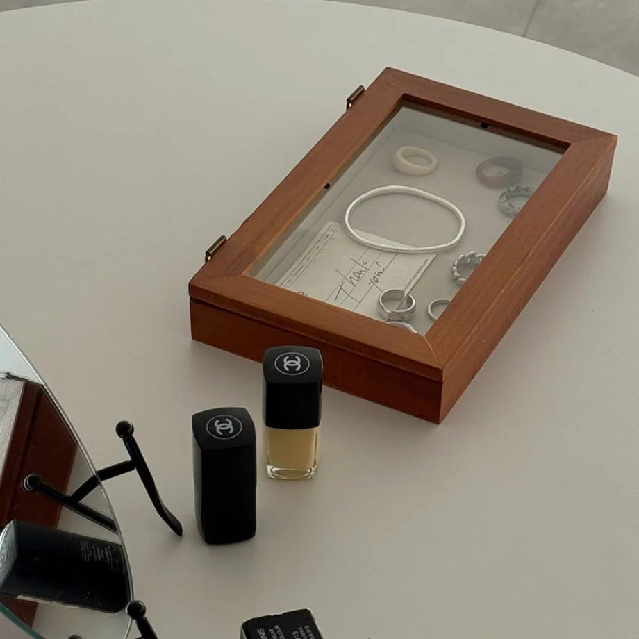 A closed wood jewelry box with a glass top sitting next to makeup on a clean white surface. - RoomDen