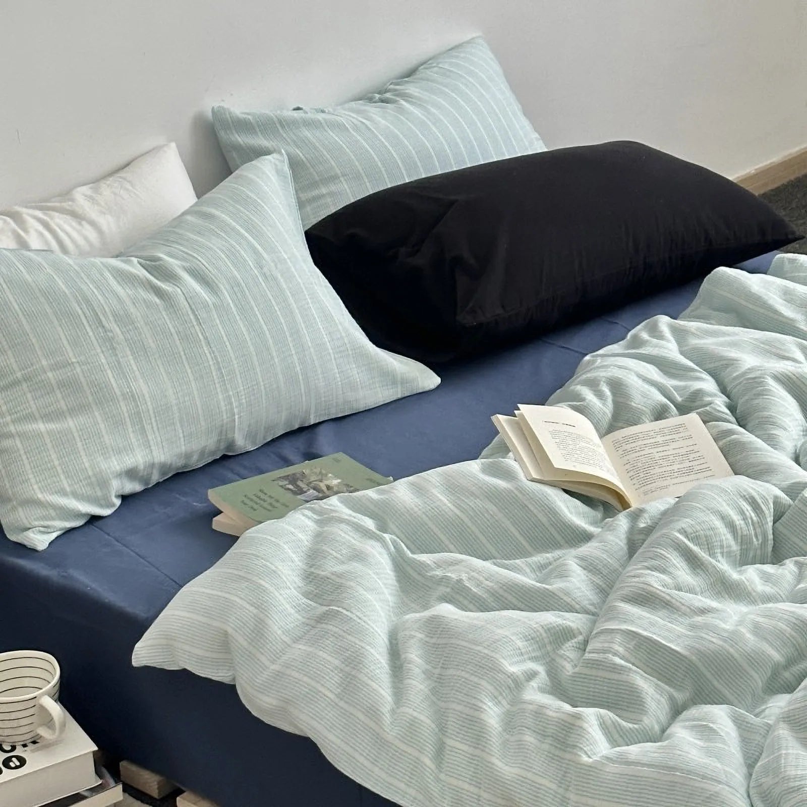 Messy bed with cyan pinstripe washed cotton duvet cover, pillows, and an open book, showcasing a relaxed vibe.