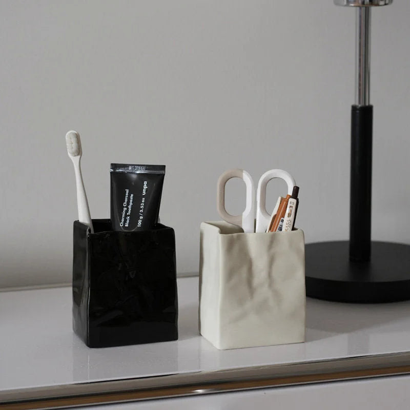 A black and a cream crumpled ceramic organizer side-by-side on a white desk, holding a toothbrush and scissors. - RoomDen