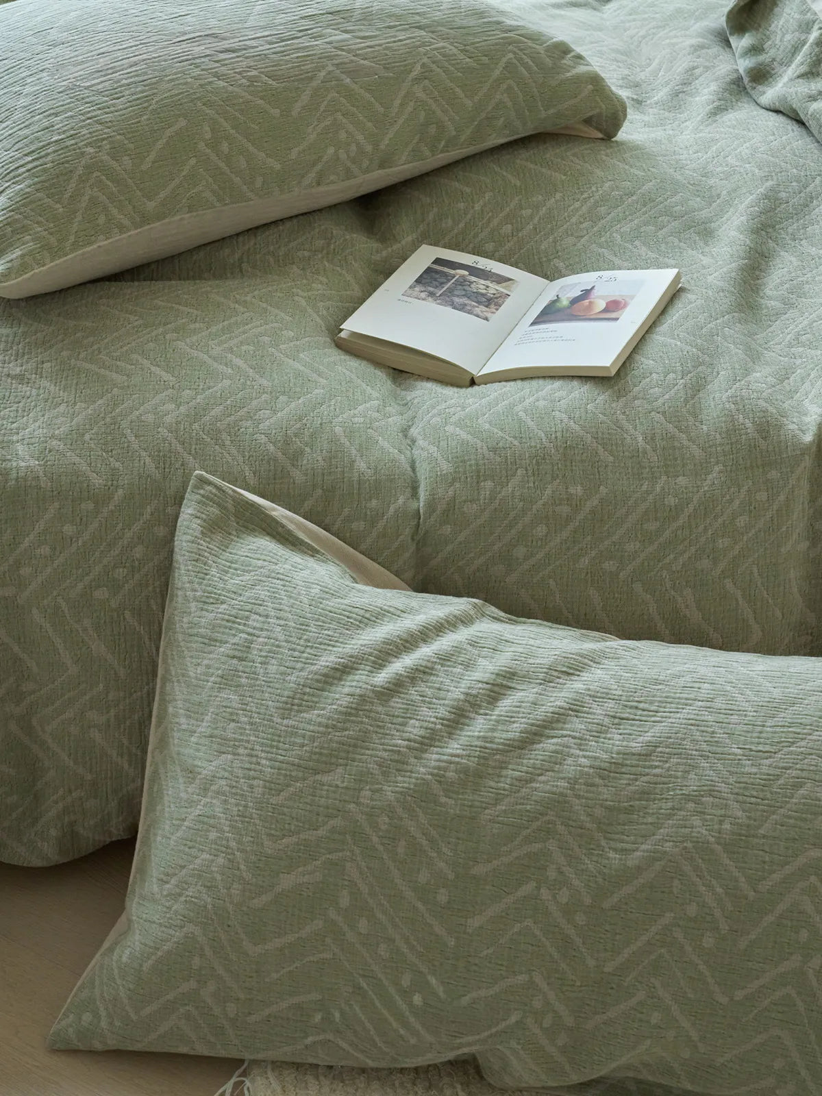 An open book resting on the sage green geometric washed cotton duvet cover, with pillows in the background. - RoomDen