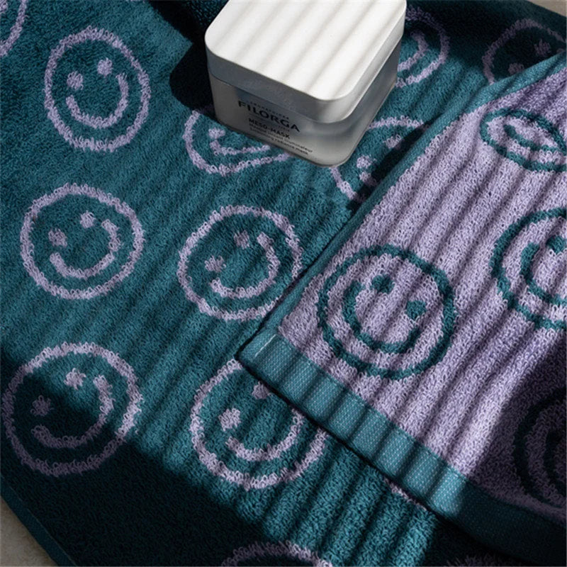 Close-up detail of the teal and lavender smiley face print on a plush cotton towel, with a skincare product resting on it - RoomDen.