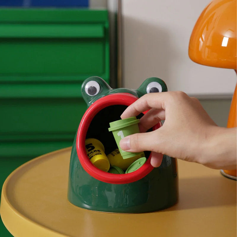 A hand placing a small item into the wide mouth of the green ceramic frog desk organizer. - RoomDen