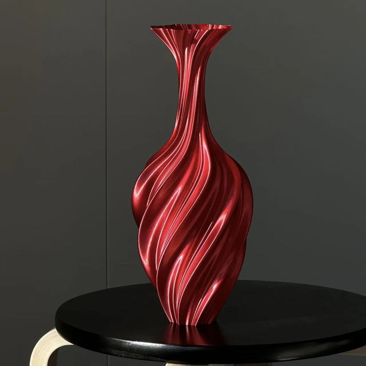 Ruby Red Sculptural Swirl Ribbed PLA Vase on a black side table against a dark wall - RoomDen