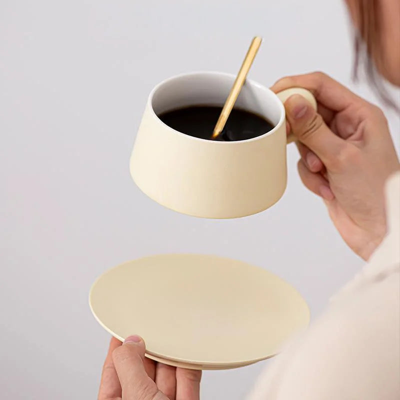 A person holding a Cream Beige matte ceramic coffee cup filled with black coffee, saucer below. - RoomDen