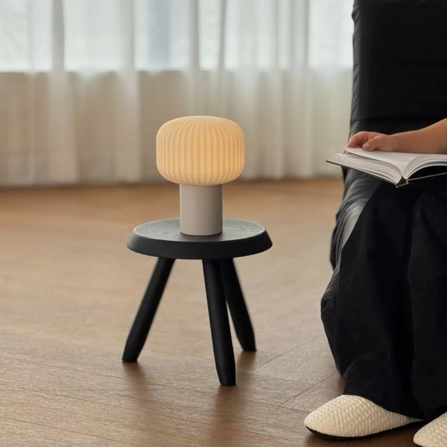 A person reading next to the glowing Pleated Glass Mushroom Accent Lamp on a stool - RoomDen