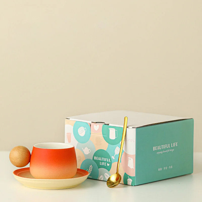 The Sunset Orange gradient ceramic mug and saucer set with a golden spoon, shown next to its stylish gift box. - RoomDen