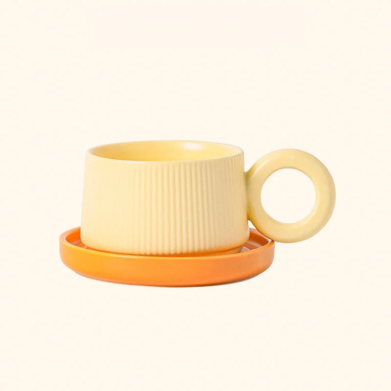 Front view of the Butter Yellow ribbed ceramic mug with its round donut handle, sitting on an orange saucer. - RoomDen