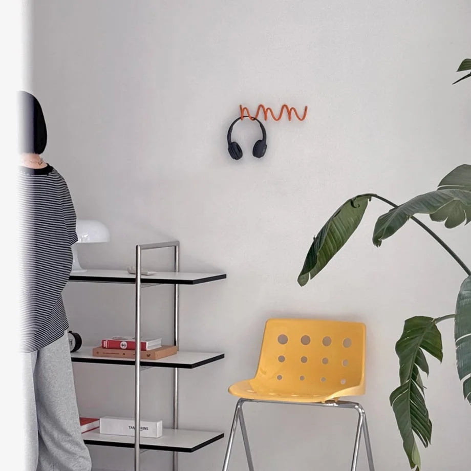 Lifestyle scene with the orange Wavy Coil Metal Wall Hook mounted in a minimalist room with a yellow chair and plant. - RoomDen