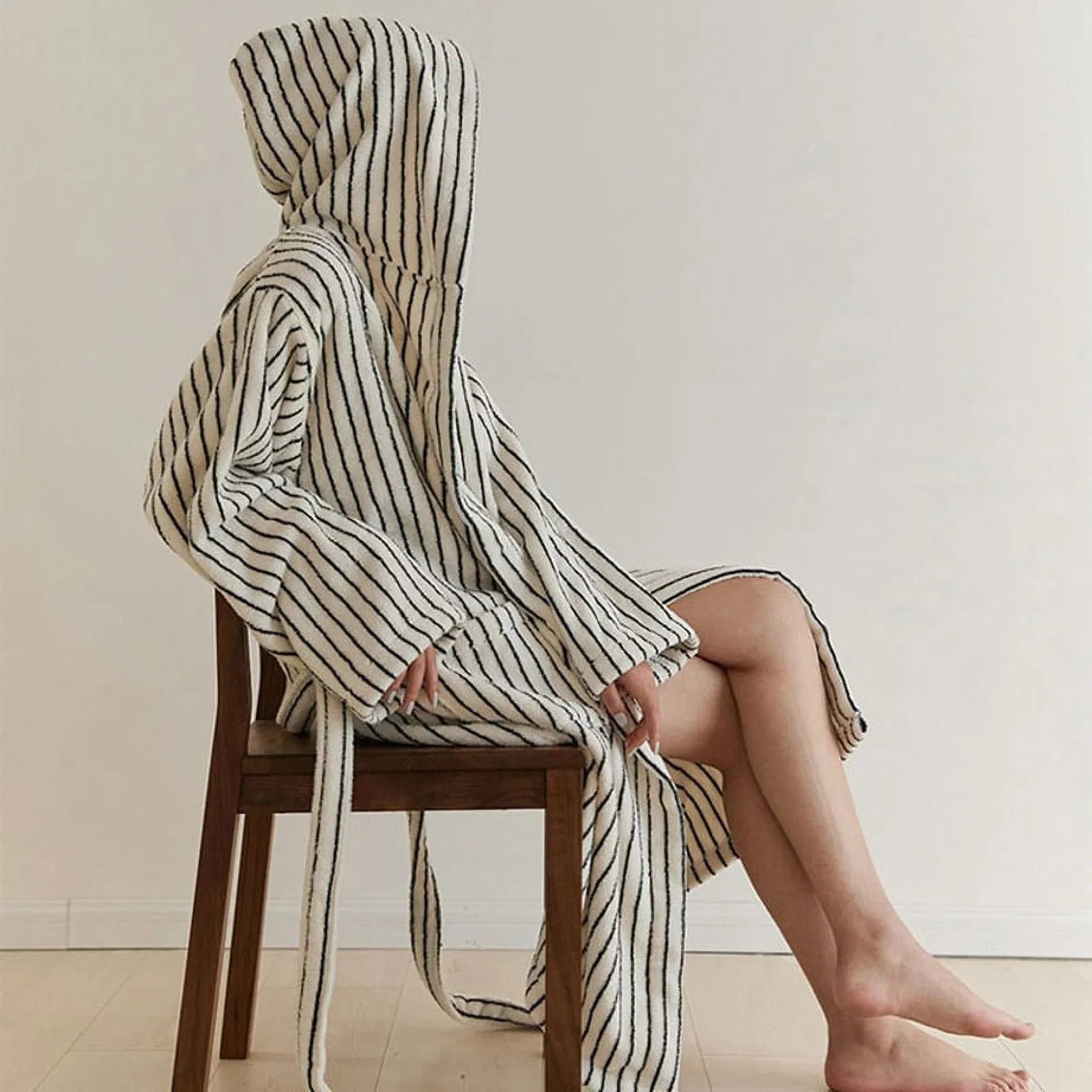 Side profile of a woman sitting on a wooden chair wearing a plush cream and black striped hooded robe.