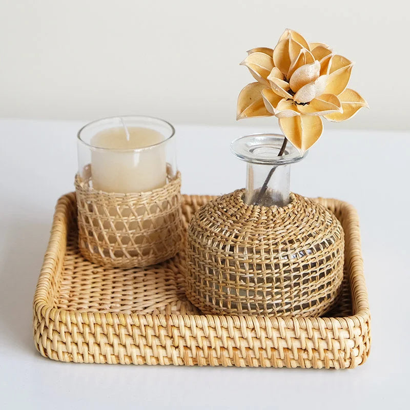 A medium Handwoven Rattan & Glass Vase used for a flower, next to a matching woven candle holder on a rattan tray.