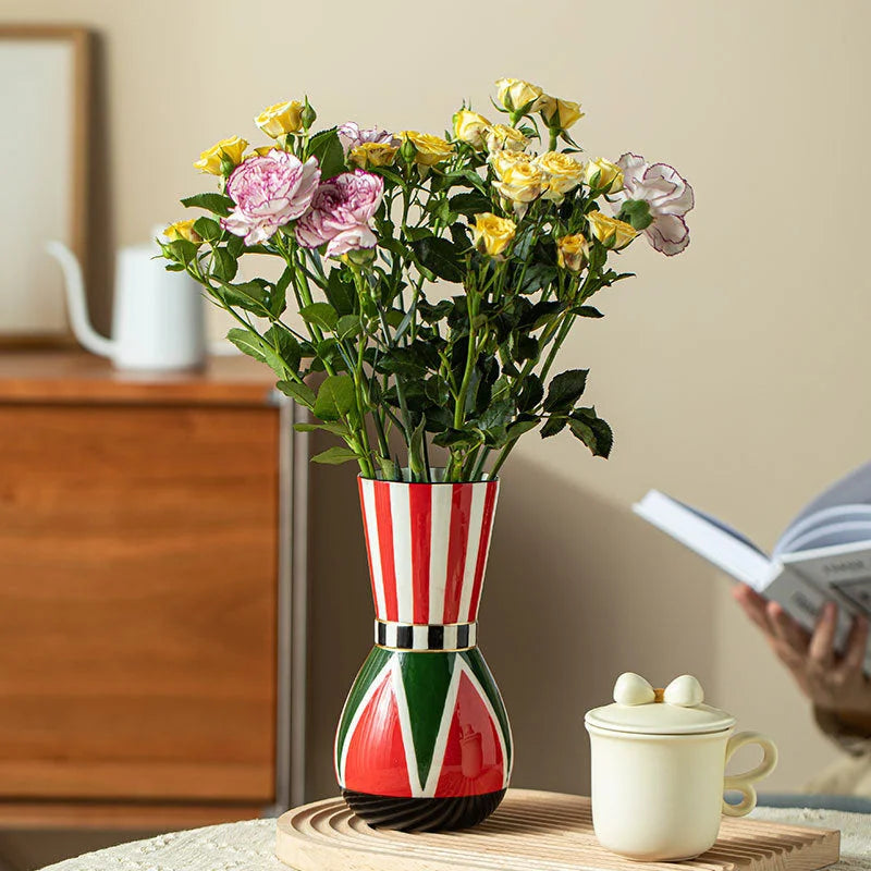 A bold red, green, and white striped ceramic vase holding a bouquet of yellow and pink roses on a wooden tray. - RoomDen