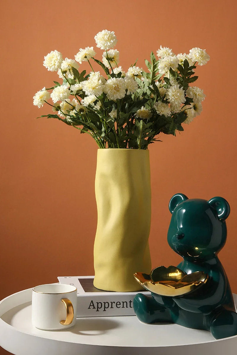 Lifestyle image showing the Lemon yellow Ruched Ceramic Vase holding white flowers on a side table next to a bear figurine - RoomDen.
