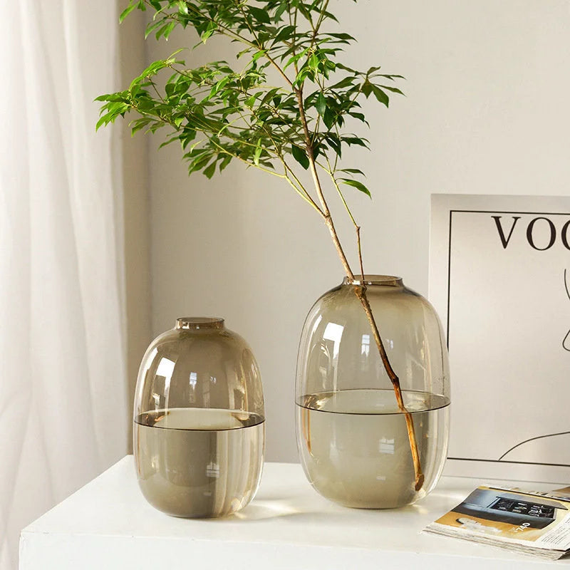 A large and small amber brown tinted glass vase on a white desk, with a green branch in the larger vase. - RoomDen