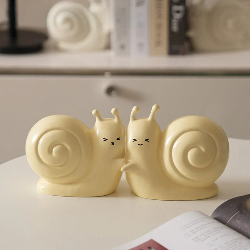 A pair of pastel yellow snail bookends cuddling together on a white tabletop, showing their adorable design - RoomDen.