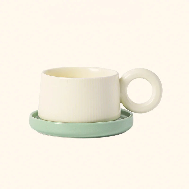 Front view of the Cream White ribbed ceramic mug with its round donut handle, sitting on a sage green saucer. - RoomDen