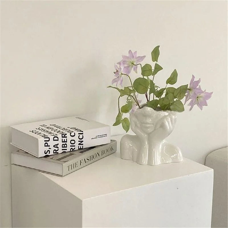 Wide shot of the white 'Daydreamer' face vase with pink flowers, next to books on a white pedestal - RoomDen.