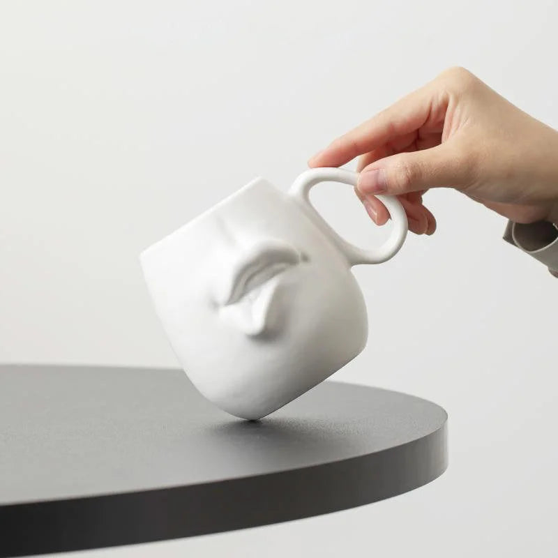 A hand holding the Matte White Sculptural Abstract Face Ceramic Mug over a black table.