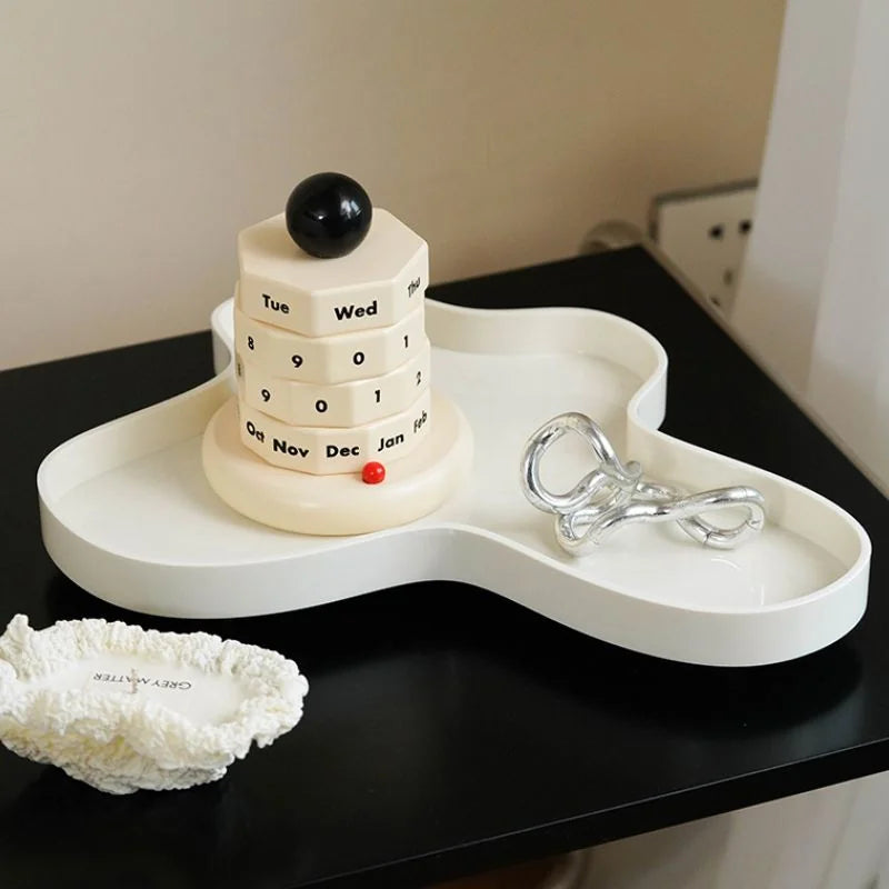The wooden perpetual calendar placed on a modern, wavy white tray next to a silver sculpture. - RoomDen