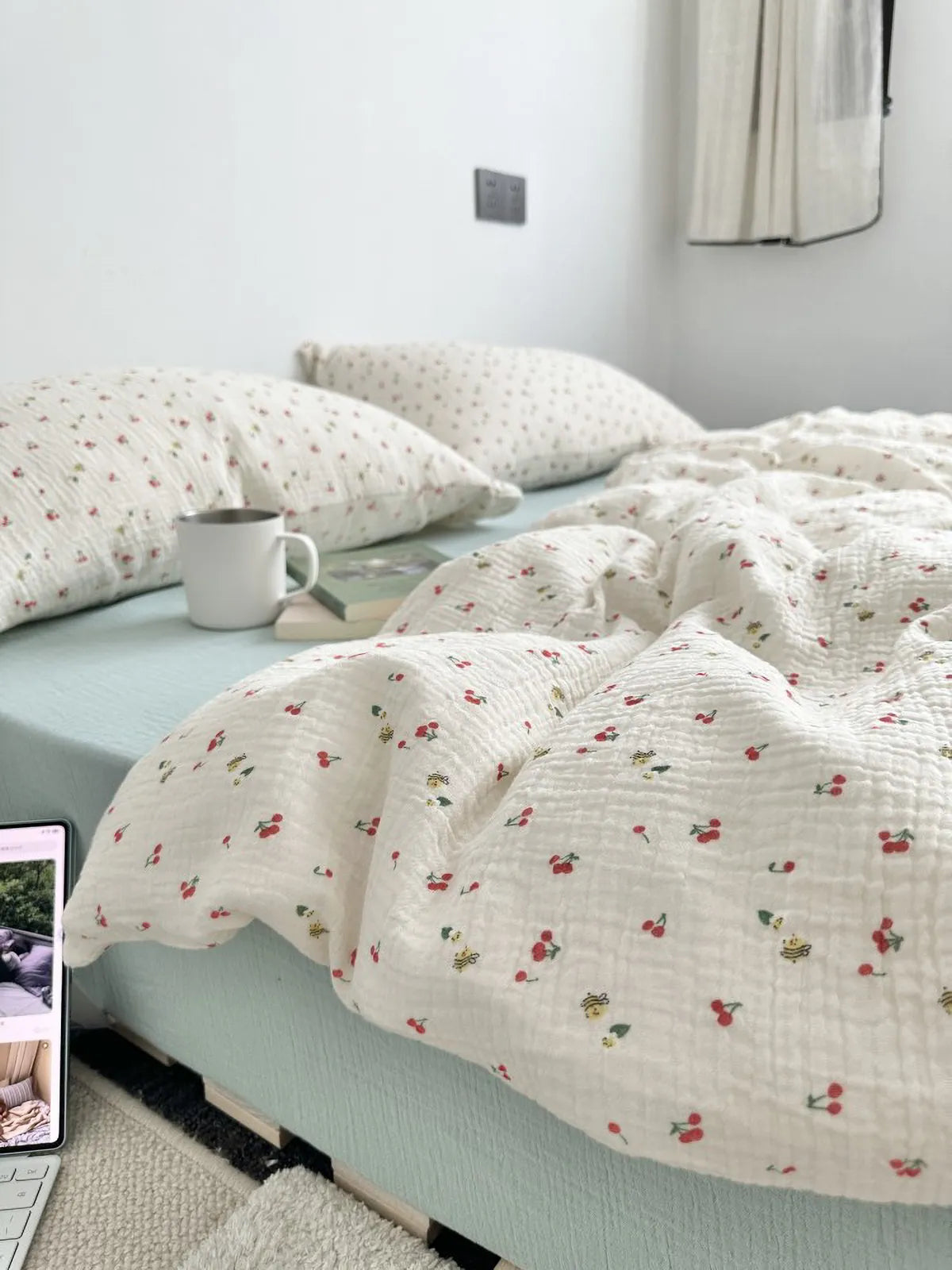 Full view of crinkled washed cotton bedding set featuring cherry and bee pattern with sage green sheets in a bright bedroom. RoomDen.