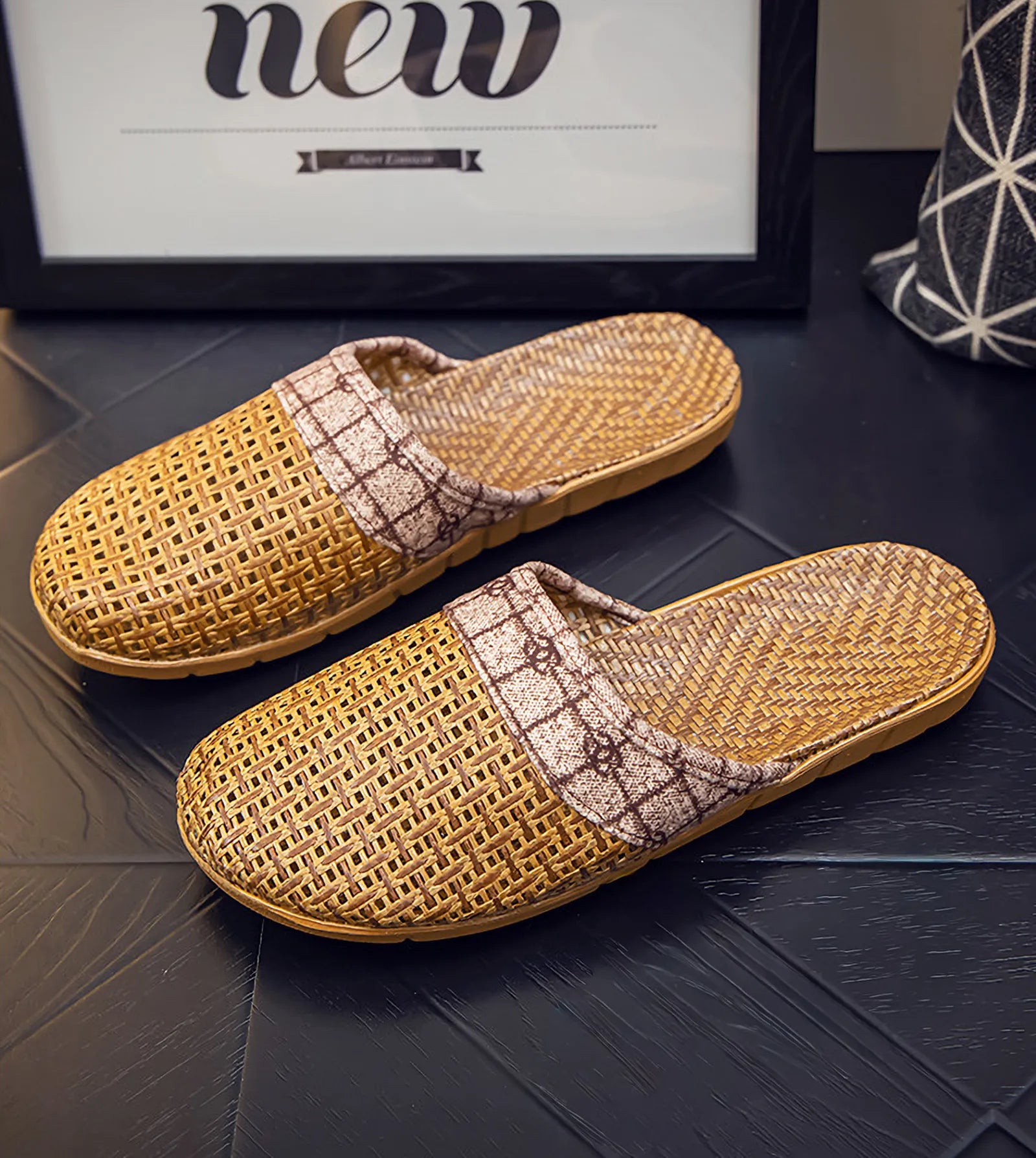 Pair of beige and brown plaid rattan slippers styled on black floor next to artwork - RoomDen