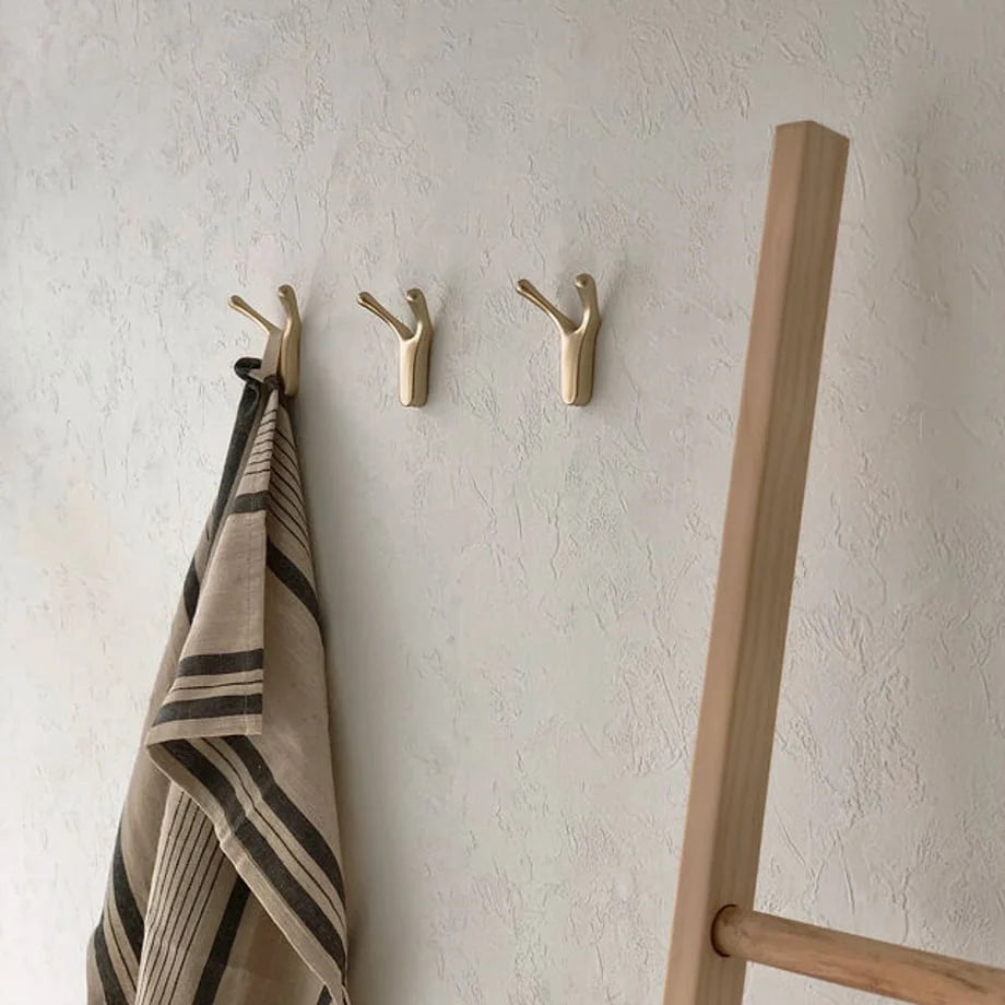 A set of three gold organic-form decorative hooks on a white wall next to a wooden ladder - RoomDen