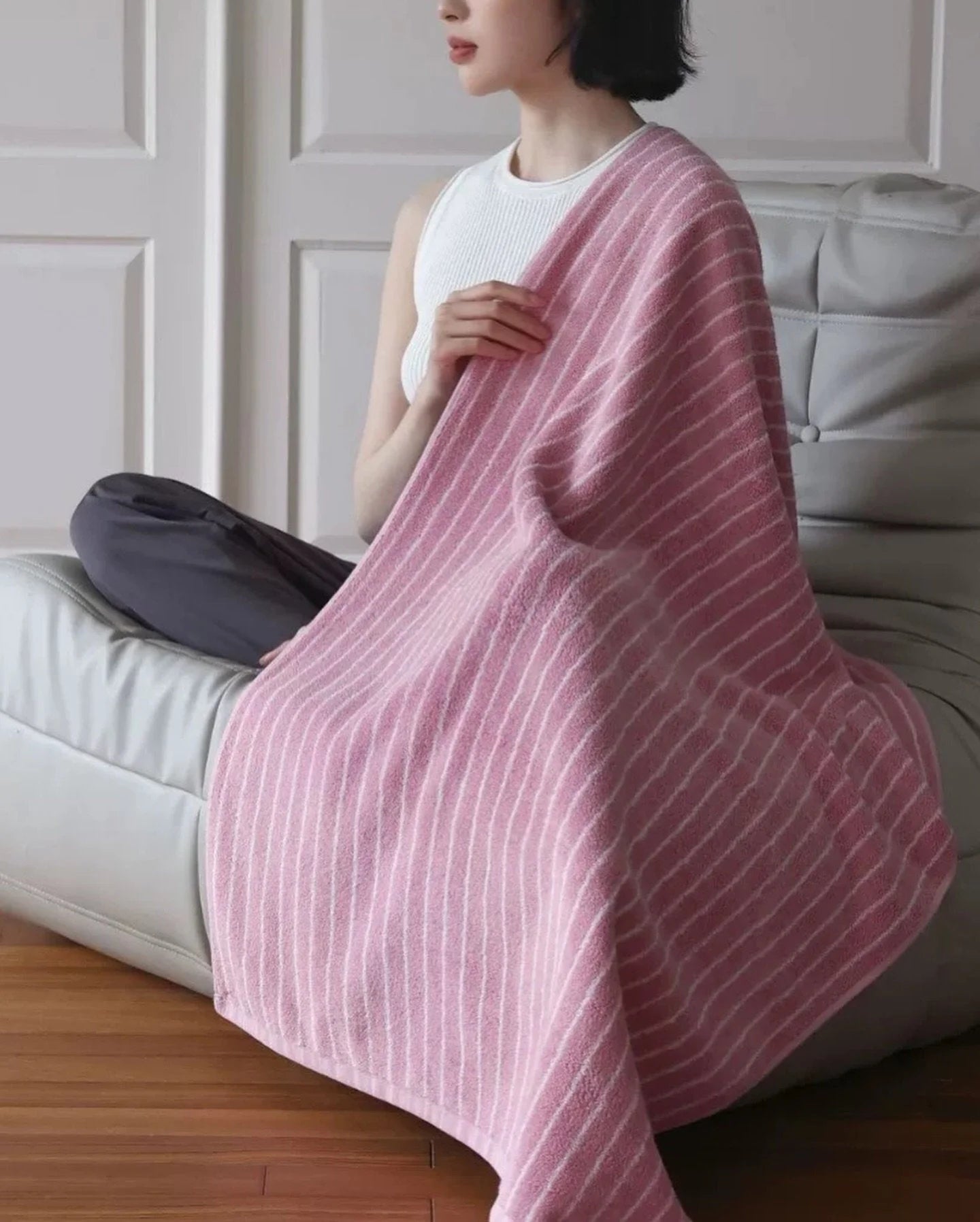 A woman sitting on a grey lounge chair, wrapped in a soft pink and white pinstripe cotton bath towel - RoomDen.