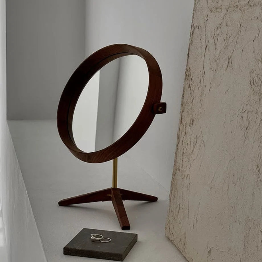 Round walnut vanity mirror with brass stand, angled view next to a textured art piece. - RoomDen
