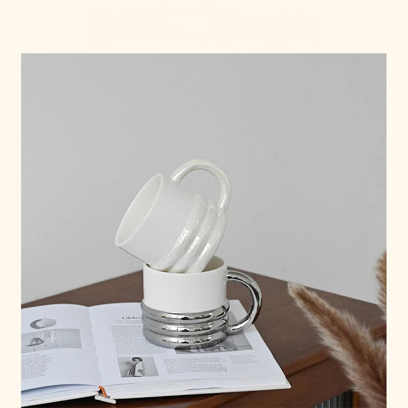Two chunky ribbed ceramic mugs, iridescent white and chrome white, stacked on a book - RoomDen.