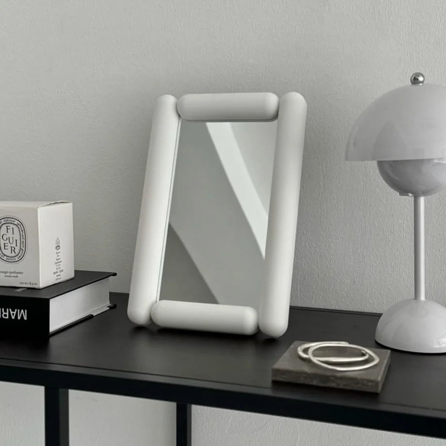 White pill-shaped desktop mirror on a dark console table next to a lamp and books. - RoomDen