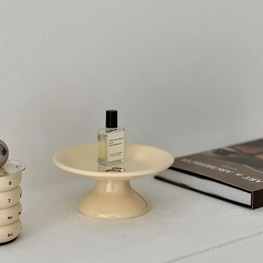 Angled view of the creamy beige pedestal trinket tray displaying a perfume bottle and rings. - RoomDen