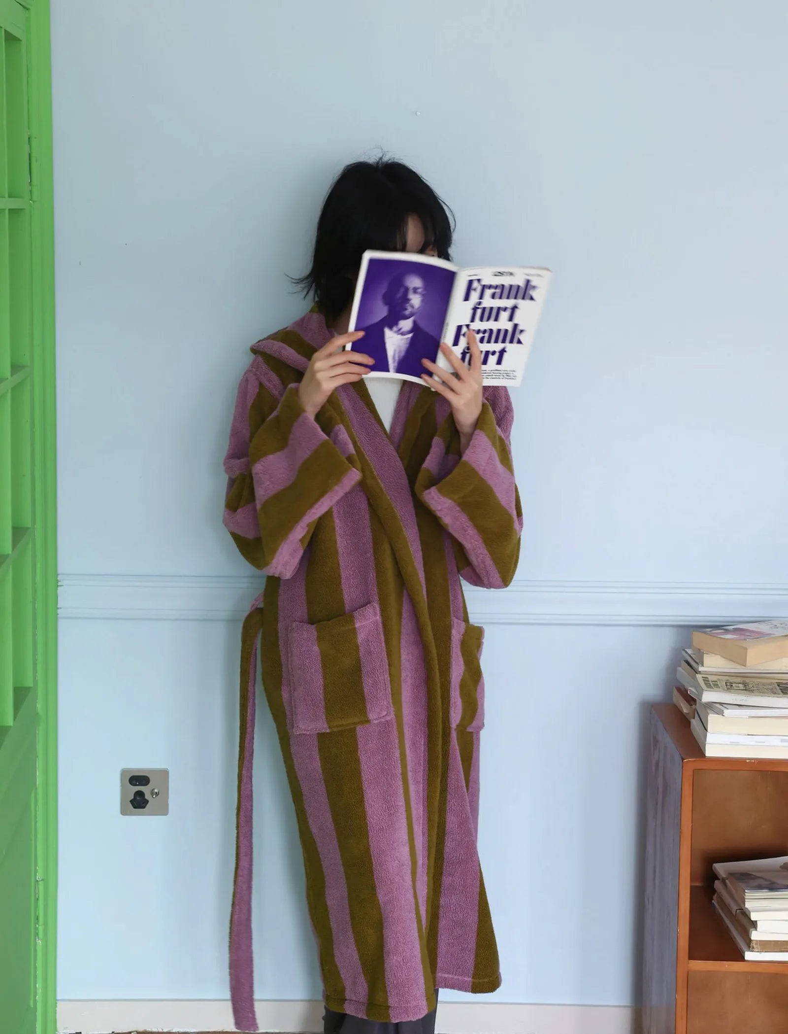 Woman holding a book covering her face wearing a purple and olive green striped hooded robe.