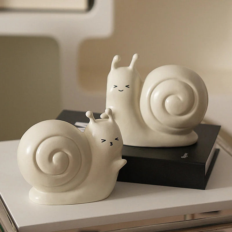 Close-up of the creamy white snail ceramic bookends, one resting on a black book, showing their cute faces - RoomDen.