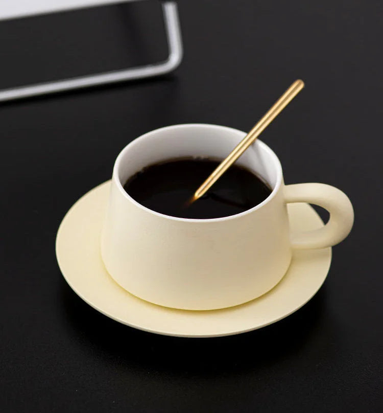 A Cream Beige matte ceramic coffee cup and saucer set with a golden spoon, on a black surface. - RoomDen