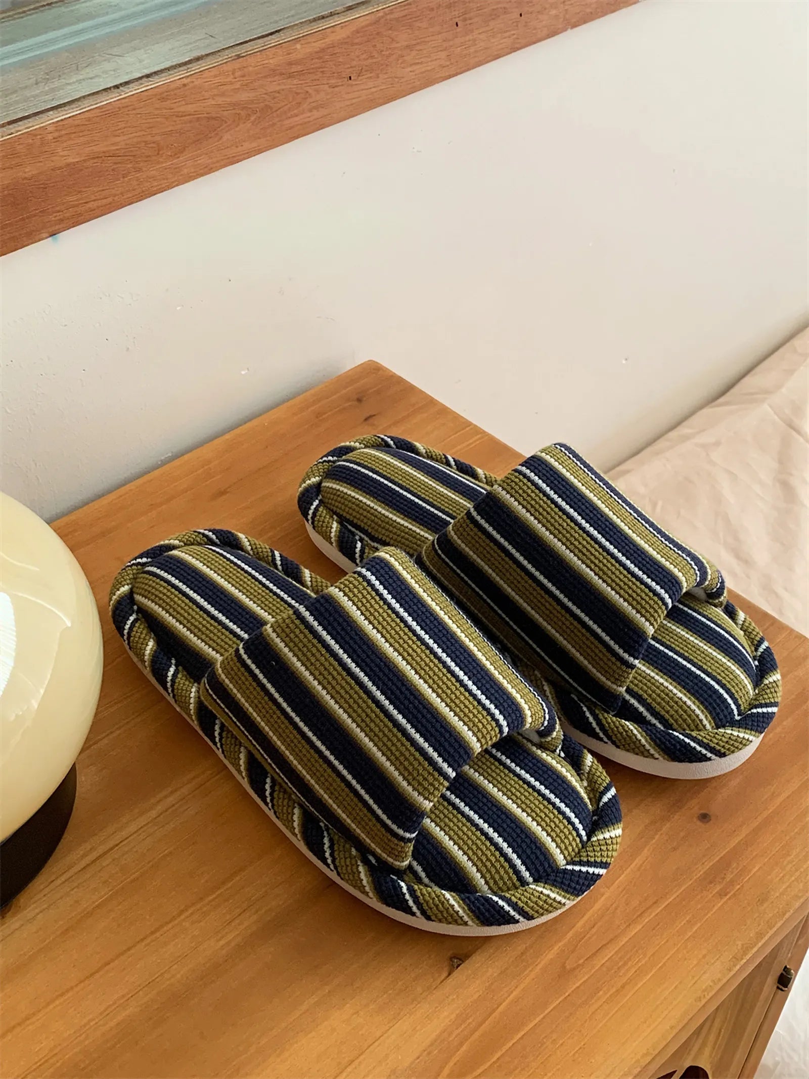 Green & White Striped and Navy & Yellow Striped slippers styled on a wooden bedside table with a modern lamp.