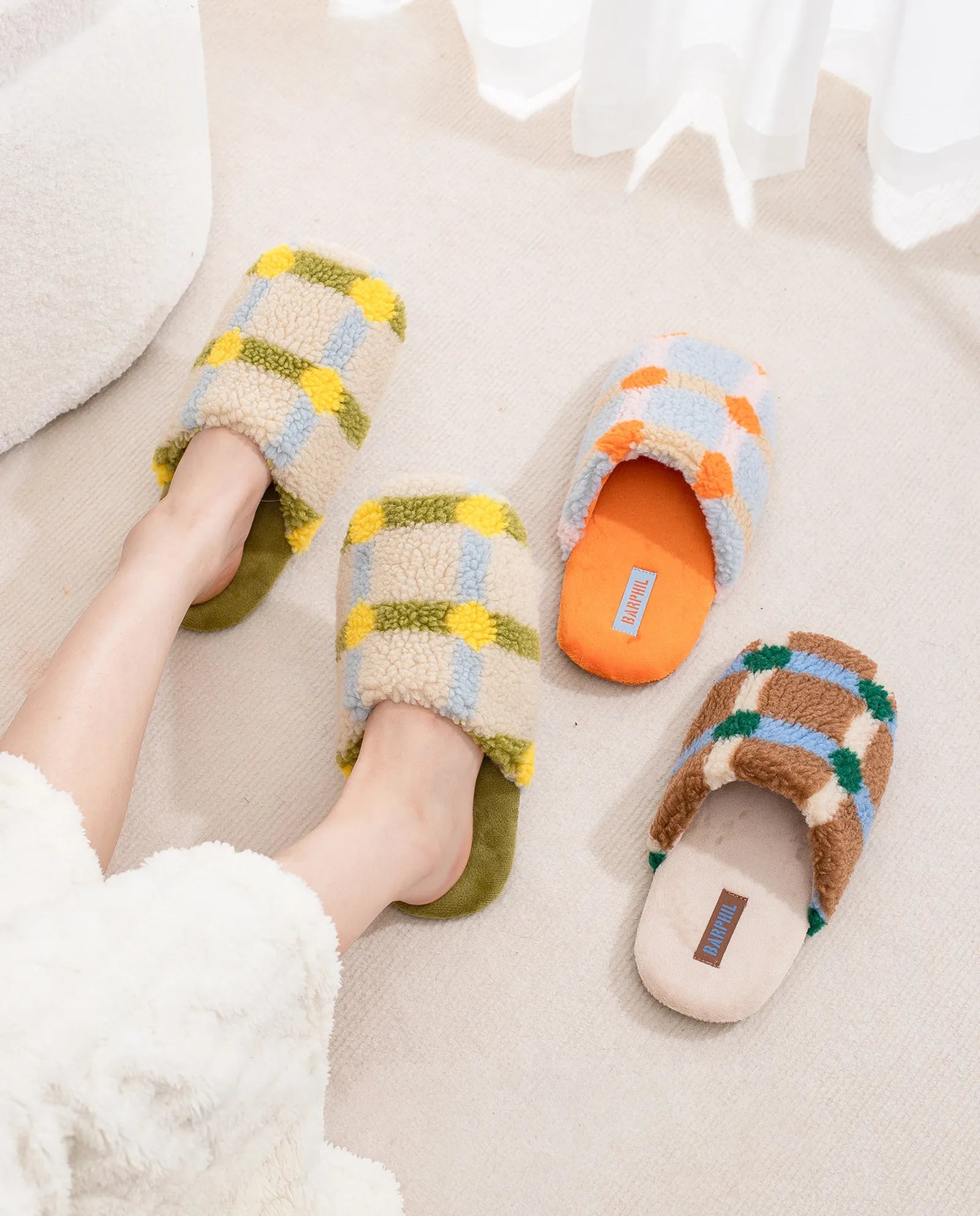 Three pairs of Fuzzy Plaid Sherpa House Slippers in Green, Orange, and Brown colorways on a rug - RoomDen