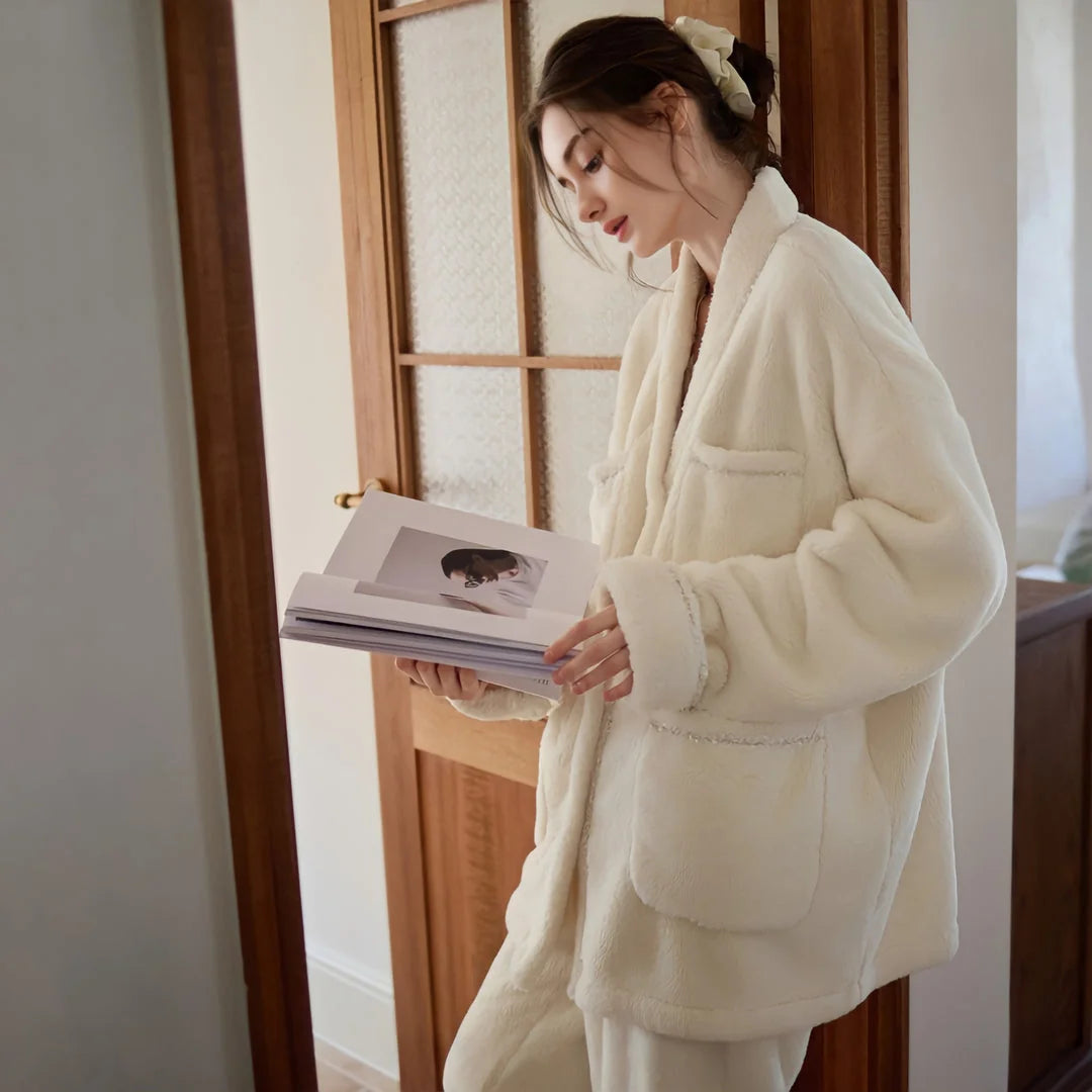 A woman in the cream fleece pajama set standing by a wooden door, looking down at a magazine in her hands - RoomDen.
