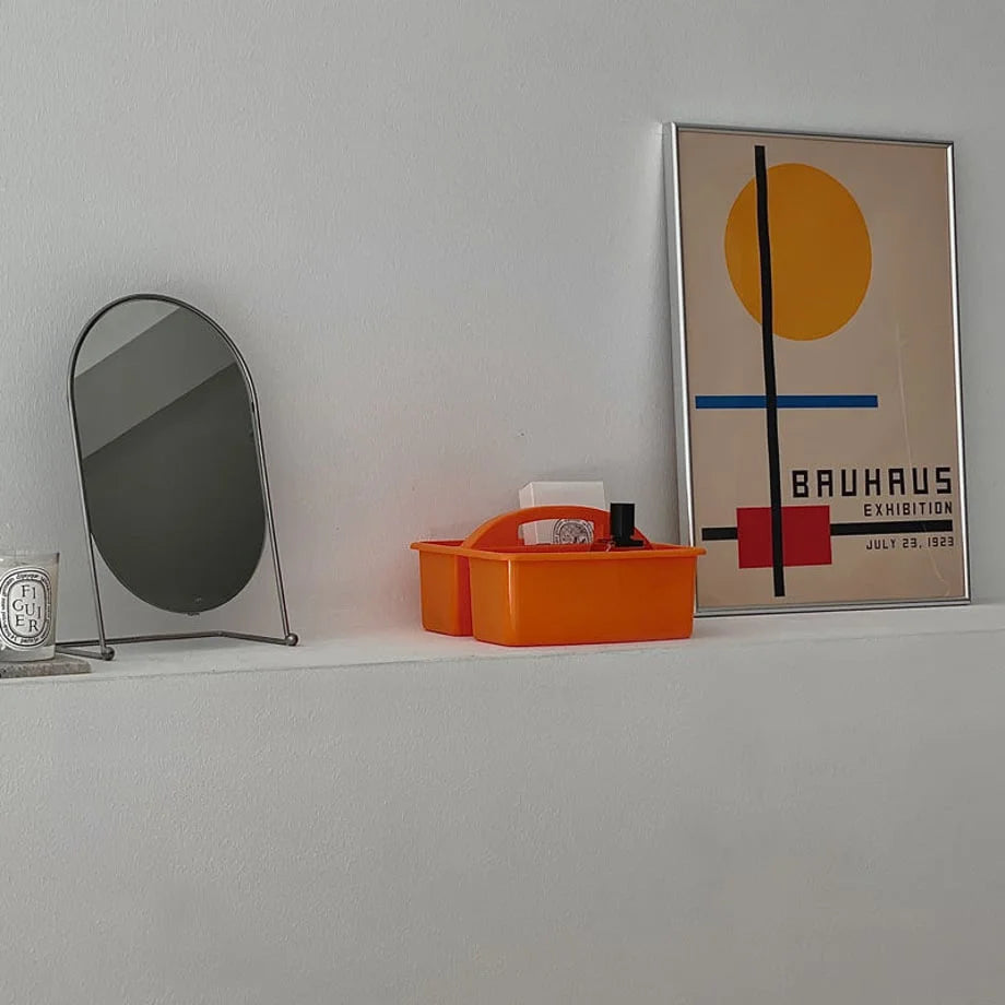 Orange portable storage caddy on a white shelf with a tabletop mirror and a Bauhaus exhibition poster. - RoomDen