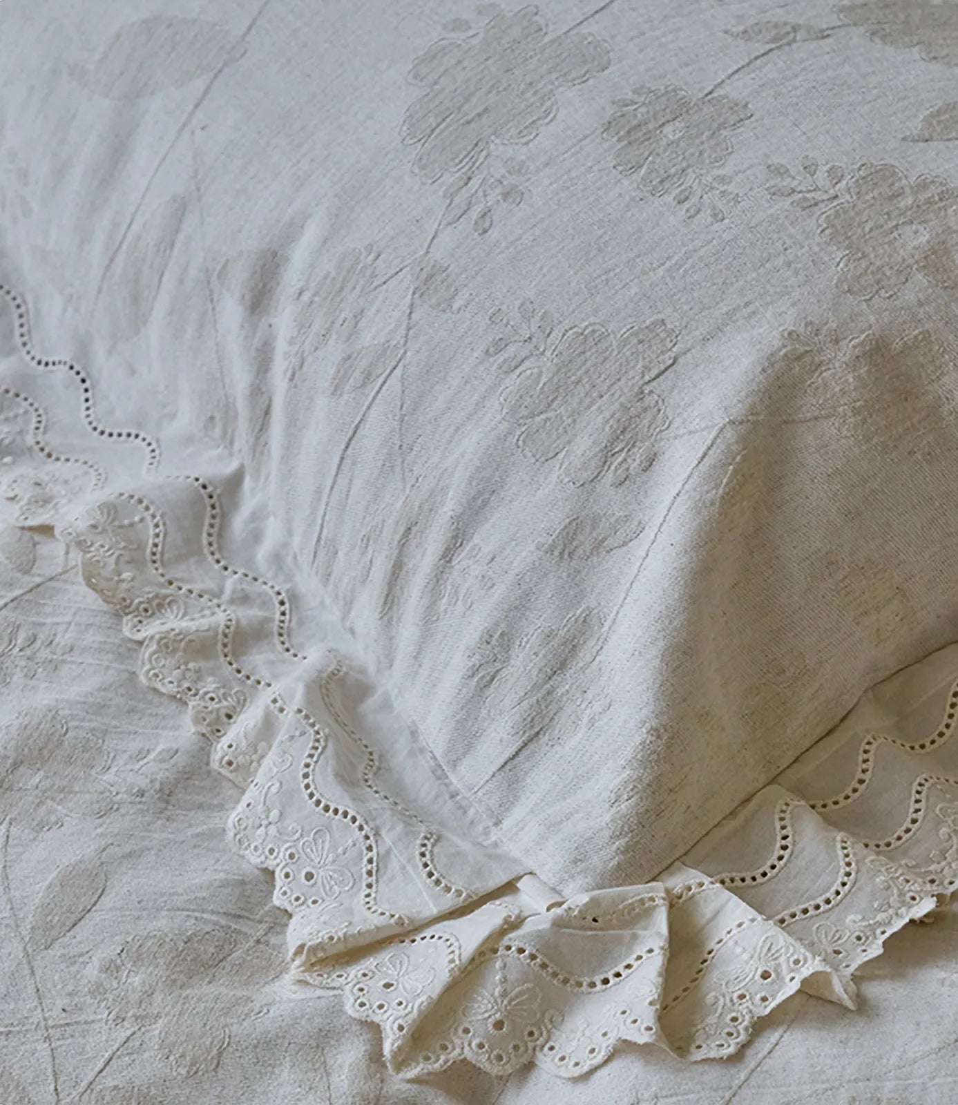 Close up of creamy linen pillowcase with floral embroidery and eyelet lace ruffle detail RoomDen.
