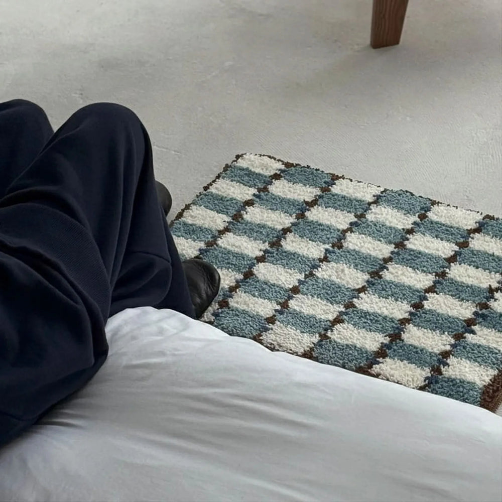 POV shot from bed looking down at Wavy Checkerboard Plush Accent Rug in teal and cream with person's legs in frame - RoomDen