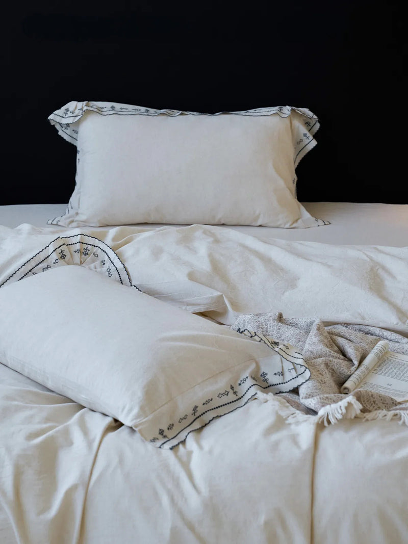 Textured Embroidered Ruffle Linen Pillowcases on a neatly made bed against a black wall, the scalloped edge reading handmade. — RoomDen